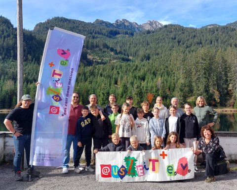 Gruppenfoto mit Menschen vor Bergen und Bäumen, Banner im Vordergrund.