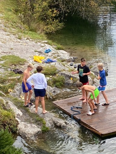 Kinder spielen am Wasser und nutzen ein Floß, mit Booten im Hintergrund.