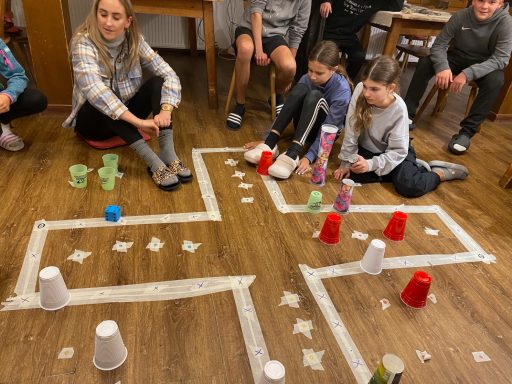 Kinder spielen auf einem Holzboden mit Bechern und einer markierten Spielfläche.